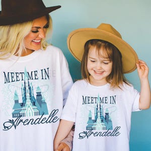 May include: White t-shirts with the text "Meet Me In Arendelle" and a blue castle graphic. The shirts are worn by a woman and a child, both smiling. They are wearing hats. The background is a solid light blue.