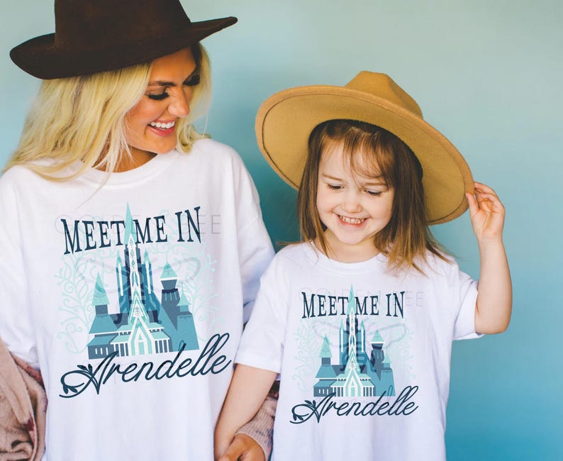 May include: White t-shirts with the text "Meet Me In Arendelle" and a blue castle graphic. The shirts are worn by a woman and a child, both smiling. They are wearing hats. The background is a solid light blue.