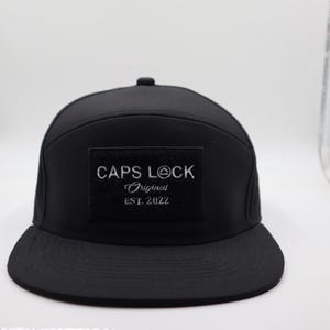 May include: A black, flat-brimmed baseball cap with a square patch on the front. The patch features the text "CAPS LOCK Original EST. 2022" in white. The cap has a structured crown and a curved brim.