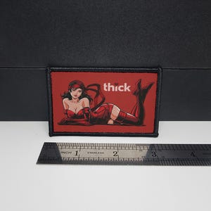 May include: Rectangular patch with a red background and a cartoon illustration of a woman in a red outfit. The word "thick" is printed in white. The patch is approximately 3 inches wide.