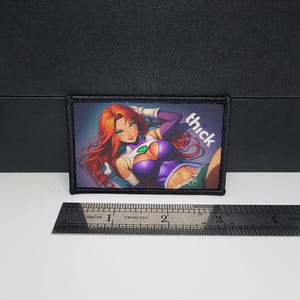 May include: A rectangular patch with an illustration of a character with red hair, green eyes, and a purple and green outfit. The word "thick" is written in white. The patch is approximately 7.6 cm long.