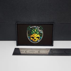 May include: Black rectangular patch featuring a yellow skull-shaped pineapple design with the word "HAWAII" above. The design includes palm trees and ocean scenery. The patch is approximately 3 inches wide.