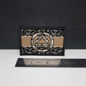 Könnte beinhalten: Ein rechteckiger Aufnäher mit einem schwarzen Paisley-Muster-Rand. Die Mitte zeigt einen horizontalen, beigen Streifen mit einem kreisförmigen Emblem, das ein Karomuster und einen nach oben zeigenden Pfeil enthält. Ein Edelstahl-Lineal ist darunter platziert.