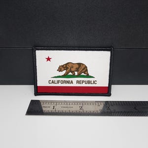 Puede incluir: Parche rectangular con la bandera del estado de California. Presenta un oso pardo, hierba verde y una estrella roja sobre un fondo blanco. La parte inferior tiene una franja roja con las palabras "CALIFORNIA REPUBLIC".