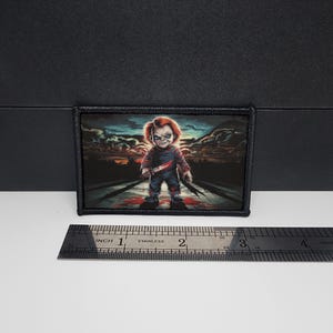 May include: A rectangular patch featuring a menacing cartoon doll with red hair, holding a knife in each hand. The doll is standing on a road with a sunset backdrop. The patch is approximately 3 inches wide.