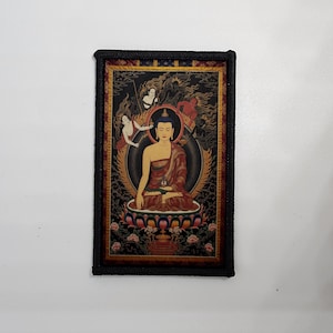 Pode incluir: Impressão de arte retangular com uma figura de Buda sentado em vestes vermelhas e douradas, rodeada por figuras e uma base floral. A obra de arte é emoldurada com uma borda preta texturizada, sobre um fundo branco.
