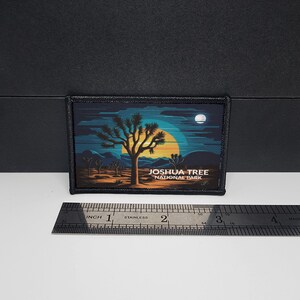 Op de afbeelding: Een rechthoekige patch met een woestijnlandschap met een Joshua-boom tegen een gele en blauwe zonsondergang. De tekst "JOSHUA TREE NATIONAL PARK" is in het wit gedrukt. De patch is ongeveer 7.6 cm breed.