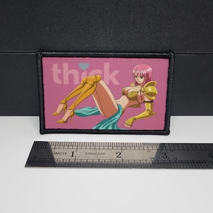 May include: A rectangular patch with a black border featuring an anime-style illustration of a woman in gold armour and a teal skirt, with the word "thick" above. The patch is approximately 7.6 cm wide.