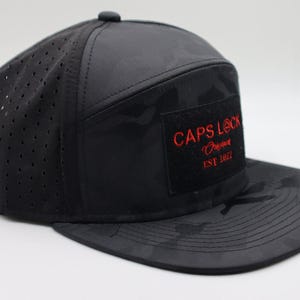 May include: Black camouflage baseball cap with a flat brim. The cap features a perforated side panel and a square patch with the red text "CAPS LOCK Original EST 2022". The cap has a button on top.