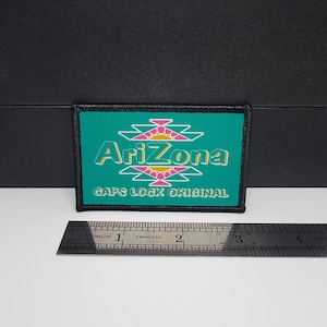 May include: Rectangular patch with the word "Arizona" in white, teal, and pink, with a geometric design above and below the text. The text "GAPS LOCK ORIGINAL" is below the word "Arizona". A ruler is placed below the patch, measuring in inches.