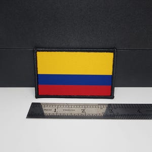 May include: A rectangular patch featuring the flag of Colombia. The flag design includes horizontal stripes of yellow, blue, and red. The patch has a black border and is placed next to a ruler showing inches.