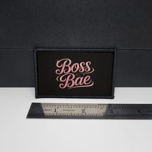 Puede incluir: Parche rectangular negro con las palabras "Boss Bae" en letras rosas y un acento dorado. El parche mide aproximadamente 7,6 cm de ancho y 3,8 cm de alto. El parche está sobre una superficie blanca.