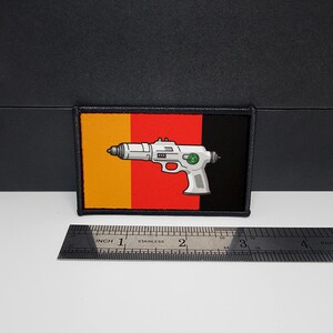 Puede incluir: Parche rectangular con un diseño de pistola de rayos blanca sobre una bandera tricolor vertical naranja, roja y negra. El parche tiene un borde negro cosido. Una regla de acero inoxidable se coloca debajo.