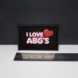 May include: A rectangular black patch with the text "I LOVE ABG'S" in white and a red heart graphic. A stainless steel ruler is placed below the patch, showing measurements in inches.