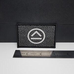 May include: Rectangular black patch with a textured surface and a silver-colored design. The design features a circle with an upward-pointing arrow and a horizontal line inside. A stainless steel ruler is placed below the patch.