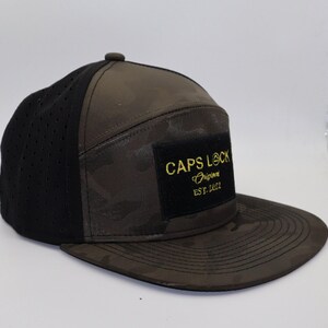 May include: A flat-brimmed baseball cap with a camouflage pattern on the crown and brim. The left side panel is black with small perforations. A black square patch on the front displays the text "CAPS LOCK Original EST. 2022" in gold.
