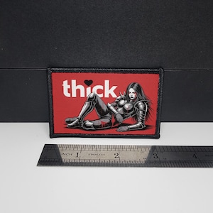 May include: A rectangular patch with a black border and a red background. The patch features the word "thick" in white with a heart symbol, and an illustration of a woman in armor. A ruler is placed below the patch.