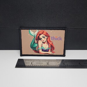 May include: A rectangular patch with a cartoon mermaid design. The mermaid has red hair and a teal tail, lying on a tan background. The word "thick" is in pink. The patch is framed by a black border.