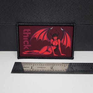 May include: Rectangular patch with a red background featuring a cartoon devil woman with red wings and horns. The word "thick" is printed vertically in white. The patch is approximately 7.6 cm long.