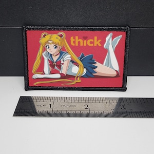 May include: A rectangular patch with a black border features an anime-style illustration of a character in a sailor uniform, lying down on a red background. The word "thick" is written in yellow. A ruler is placed below the patch.