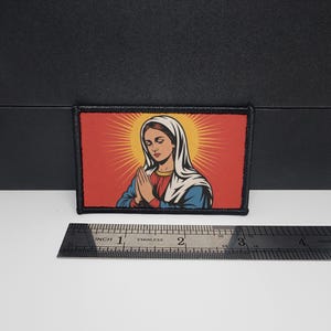 May include: Rectangular patch featuring a religious illustration. The image depicts a woman with a white veil, hands clasped in prayer, set against a red background with a yellow sunburst design. The patch is approximately 3 inches wide.