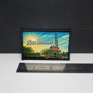 May include: Embroidered patch featuring a scenic illustration of San Leandro, California, with the Manor Shopping Center sign. The design includes a bright sun, trees, and the text "San Leandro" and "Manor Shopping Center". The patch is approximately 2 inches wide.