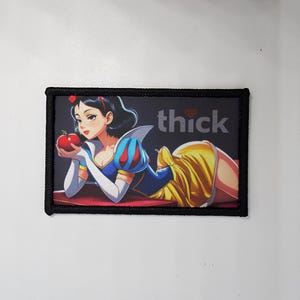 May include: A rectangular patch with a black border. It features an anime-style illustration of a woman in a blue and yellow dress holding a red apple, with the word "thick" in white.