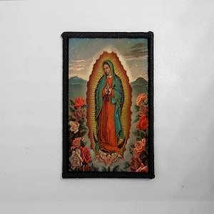 May include: Rectangular patch featuring an image of Our Lady of Guadalupe. The image shows a woman in a red and teal robe, surrounded by a golden aura, roses, and mountains. The patch has a black border.