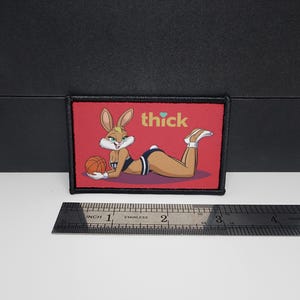 May include: A rectangular patch with a cartoon bunny in a swimsuit, holding a basketball, and the word "thick" above. The patch has a red background and a black border. A ruler is placed below the patch.