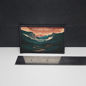Könnte beinhalten: Rechteckiger Aufnäher mit einer Illustration des Great Smoky Mountains National Park. Das Kunstwerk zeigt einen Fluss, der sich durch ein Tal schlängelt, mit Bergen und einer Hütte. Der Aufnäher ist etwa 7.6 cm breit.