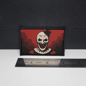 May include: A rectangular patch featuring a sinister clown with a bald head, a small black top hat, and a wide, unsettling grin. The background is red with dark, smoky clouds. The patch has a black border.