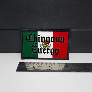 May include: Rectangular patch featuring the Mexican flag design with the words "Chingona Energy" in black gothic font. The patch is bordered in black and includes the Mexican coat of arms. A stainless steel ruler is placed below the patch.