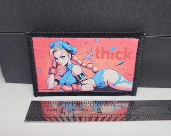 May include: A rectangular patch featuring a stylized illustration of a woman in a blue outfit and hat, with the word "thick" in pink. The patch is bordered in black and includes a ruler for scale.