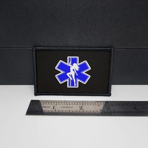 Peut inclure: Écusson rectangulaire noir avec un emblème bleu et blanc. L'emblème représente une étoile de vie bleue avec la silhouette blanche d'une femme dansant sur une barre. Une règle en acier inoxydable est en dessous.