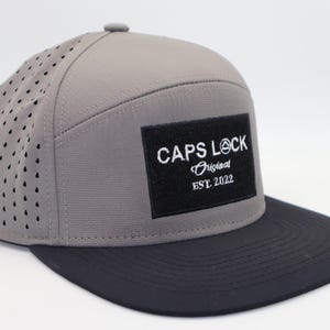 Puede incluir: Gorra de béisbol gris y negra con el texto "CAPS LOCK Original EST. 2022" en un parche cuadrado negro. La gorra tiene un panel lateral perforado y una visera plana.