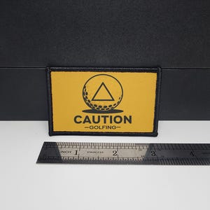 May include: A rectangular yellow patch with black trim and the words "CAUTION -GOLFING-". The patch features a golf ball with a triangle inside a circle. A stainless steel ruler is placed below the patch.