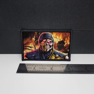 May include: Rectangular patch featuring a character with a skull face, red eyes, and a yellow and black mask. The character is set against a fiery background. The patch is approximately 3 inches wide.