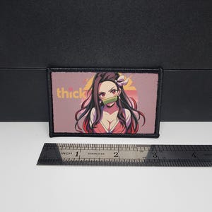 May include: Rectangular patch with an anime-style illustration of a female character with long dark hair and a bamboo muzzle. The patch has the word "thick" in yellow letters. The patch is approximately 7.6 cm long.