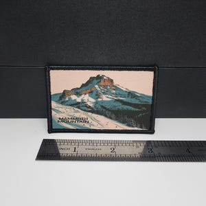 May include: Rectangular patch with a mountain scene. The patch features a snow-covered mountain with skiers and trees. The text "Mammoth Mountain" is printed at the bottom. The patch is bordered with black stitching and measures approximately 3 inches wide.