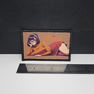 May include: A rectangular patch with an anime-style illustration of a woman in an orange turtleneck and red dress, wearing glasses. The word "thick" is above the woman, with a heart symbol. The patch is bordered in black.