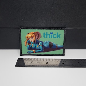 May include: A rectangular patch with a green background featuring an anime-style illustration of a woman in a blue and black bodysuit. The word "thick" is printed in white. The patch is approximately 3 inches wide.