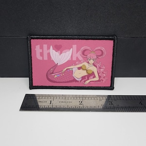 May include: A rectangular pink patch with a black border. The patch features a mermaid illustration and the word "thank" in white. The mermaid has pink hair and a pink and purple tail. A ruler is below the patch.