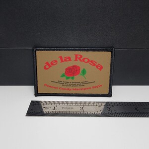 May include: A rectangular patch with the text "de la Rosa" in red, a red rose graphic, and the text "Life is like a peanut candy. sometimes it's sweet and sometimes it's just plain nuts. Peanut Candy Marzipan Style." A ruler is below the patch.