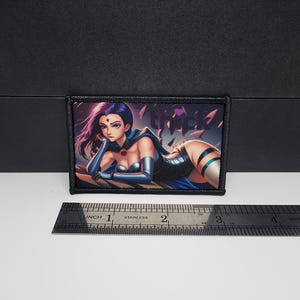 May include: Rectangular patch with an anime-style illustration of a character with purple hair, blue eyes, and a black and blue outfit. The patch has a black border and the word "thick" is visible. A ruler is placed below the patch.