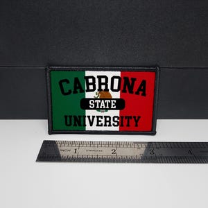 May include: Rectangular patch with the text "CABRONA UNIVERSITY" in black letters over a green, white, and red background, resembling a flag. The patch includes the words "STATE" and a brown bear graphic. A stainless steel ruler is placed below the patch.