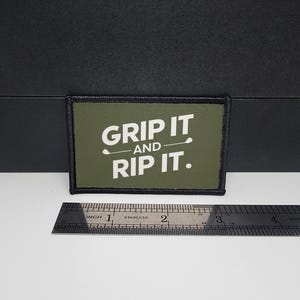 May include: An olive green rectangular patch with the text "GRIP IT AND RIP IT" in white. A golf club graphic is between the words. The patch is approximately 7.6 cm long.
