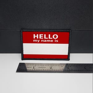 以下が含まれることがあります： 赤い上部と白い下部を持つ長方形のパッチ。「HELLO my name is」のテキストが赤い部分に白で印刷されています。ステンレス鋼の定規が下に配置されています。