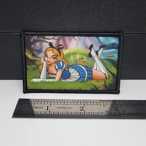 May include: A rectangular patch with a cartoon illustration of a woman in a blue and white dress, lying on grass. The patch has a black border and the text "thick" is visible. A ruler shows the patch is approximately 7.6 cm long.