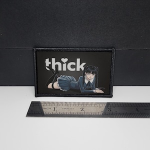 May include: A black rectangular patch with the word "thick" in white. It features an anime-style illustration of a person in a blue dress and black stockings. A stainless steel ruler is placed below the patch.
