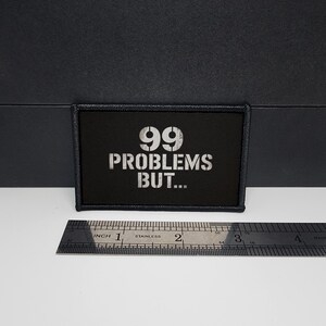 Könnte beinhalten: Schwarzer rechteckiger Aufnäher mit dem Text "99 PROBLEMS BUT..." in weißer Schablonenschrift. Der Aufnäher ist ca. 7,6 cm lang und 3,8 cm breit. Der Aufnäher hat einen schwarzen Rand.